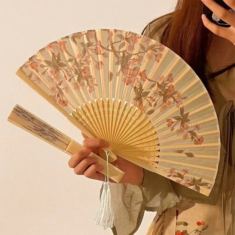 A hand holds an elegant floral bird print bamboo folding fan with light wood and pink accents.