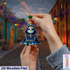 A hand holds a blue and teal flat wooden skull ornament decorated with flowers, set against a backdrop of an old-style street