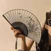 A hand holds an elegant floral bird print bamboo folding fan with a black frame and striped design.