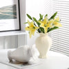 A white ceramic vase holds yellow lilies against a bright background.