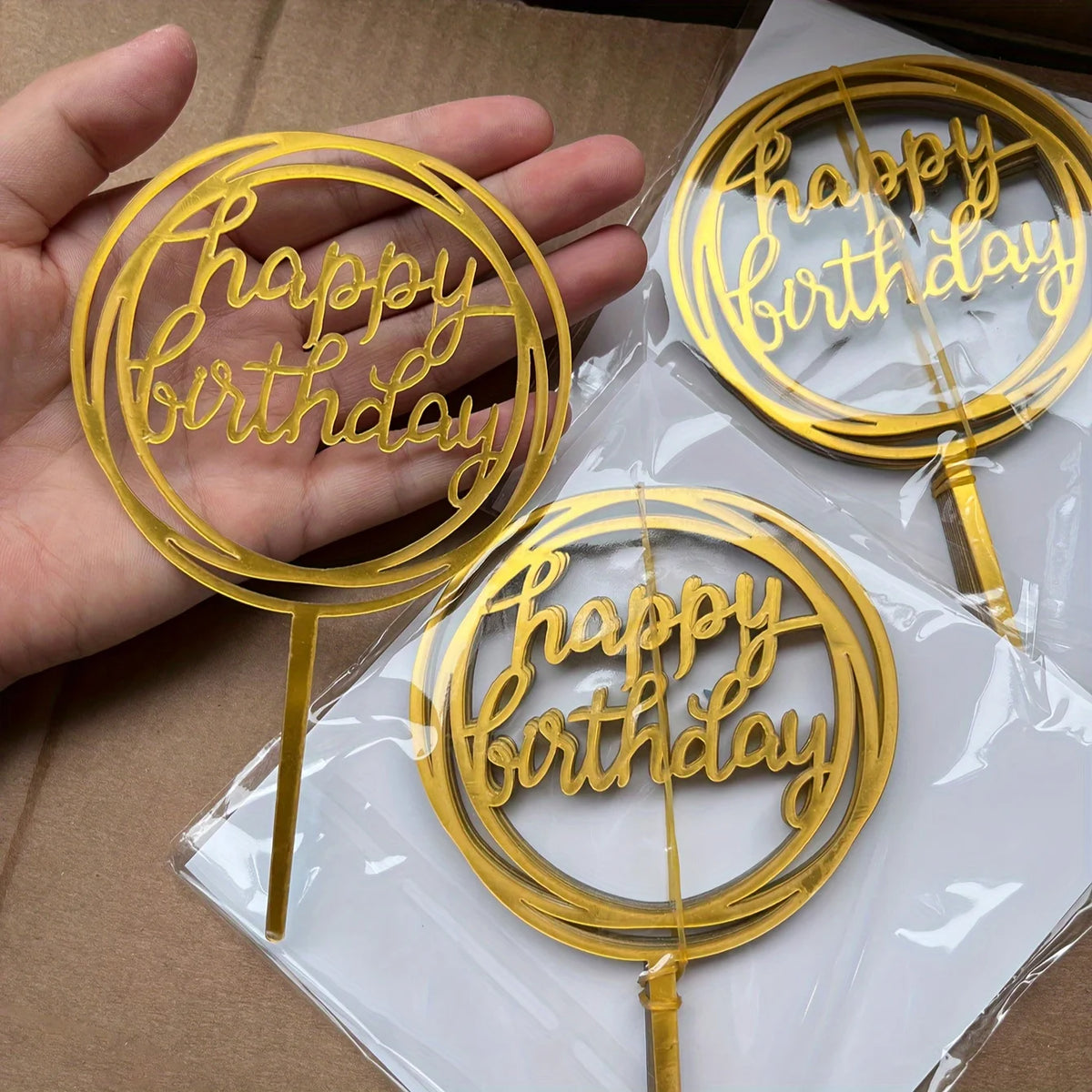 A set of ten gold acrylic birthday cake toppers with the words Happy Birthday on them, displayed in clear plastic packaging.