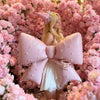 A blonde woman wearing a pink dress holds a large pink pearl bow balloon weight surrounded by pink floral decorations.