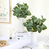 A white ceramic vase holds a lush green artificial eucalyptus stem arrangement on a white table.