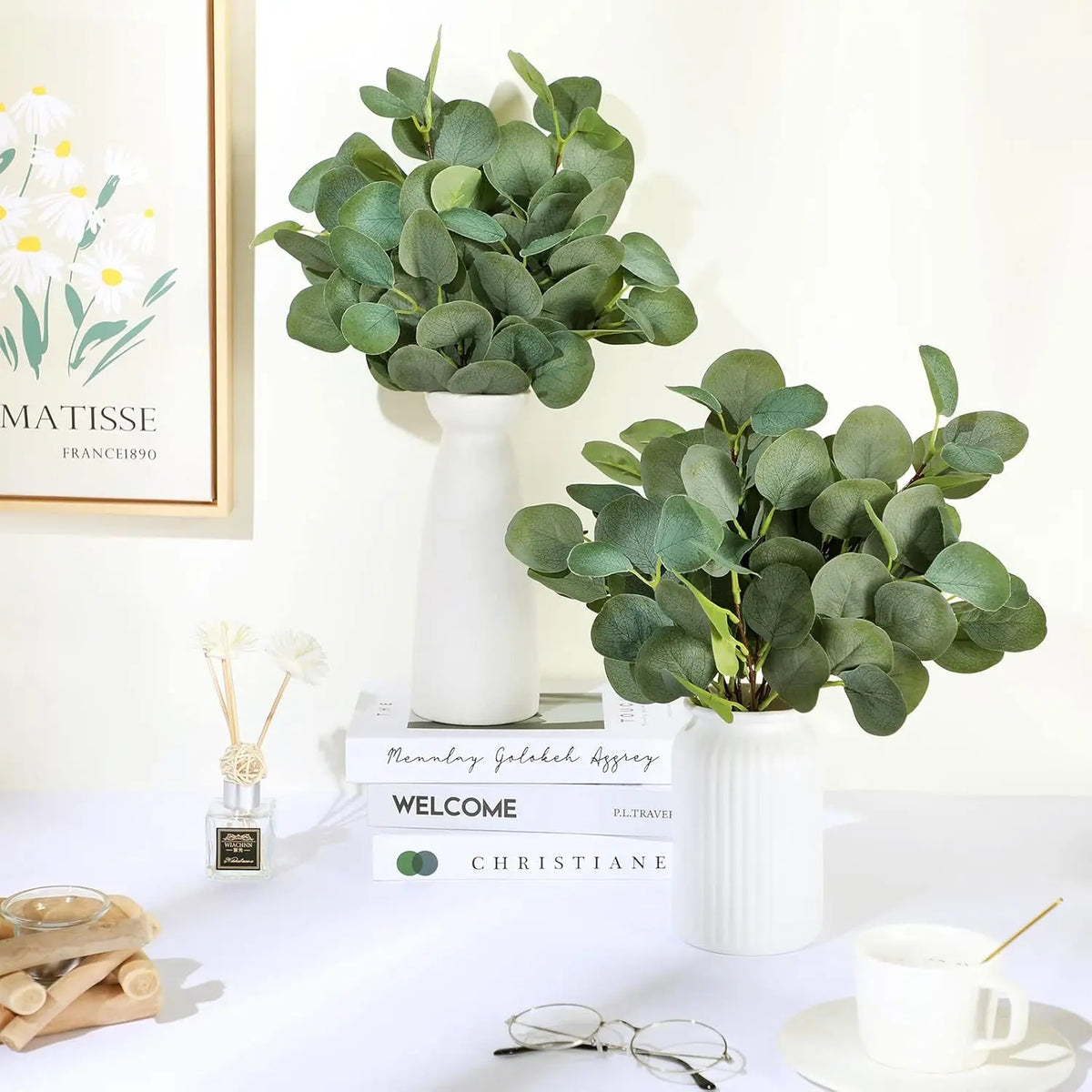 A white ceramic vase holds a lush green artificial eucalyptus stem arrangement on a white table.