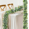 A long, lush green artificial eucalyptus vine garland decorated with white flowers on a draped table setting.