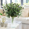 A vase filled with artificial olive leaf branches on a white sofa with light-colored pillows.