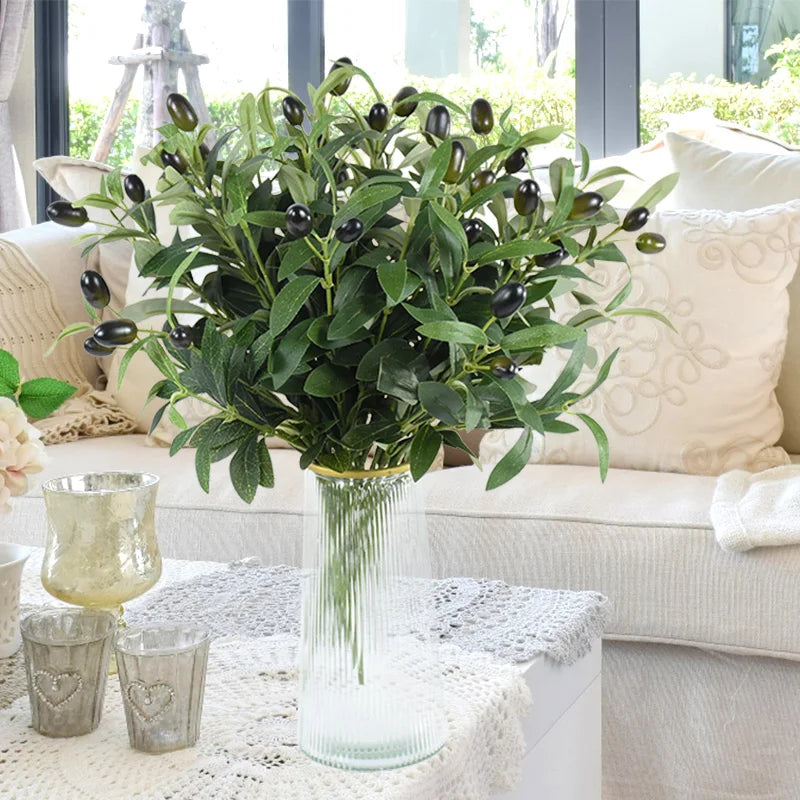 A vase filled with artificial olive leaf branches on a white sofa with light-colored pillows.