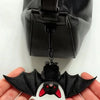 A black plush bat keychain with red accents is held in a hand against a dark leather bag.