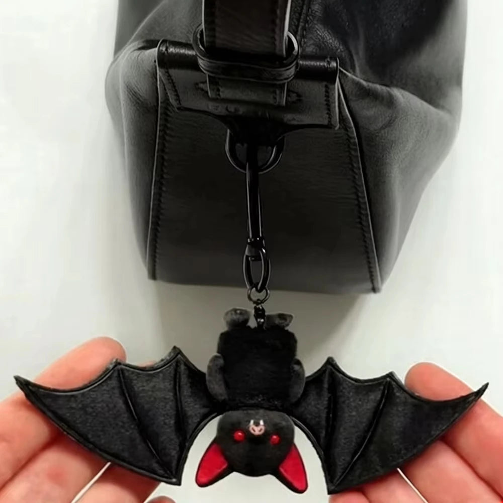 A black plush bat keychain with red accents is held in a hand against a dark leather bag.