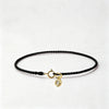 A black string bracelet with a gold plated clasp and a round gold charm on a white background.