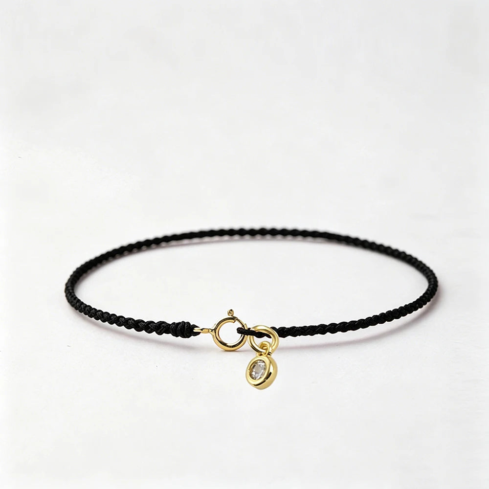 A black string bracelet with a gold plated clasp and a round gold charm on a white background.