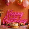 A pink neon wall sign spelling out Happy Birthday with balloons and floral arrangements in an orange setting.