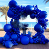 A large balloon arch garland made of royal blue balloons decorated with palm fronds on a wooden deck.