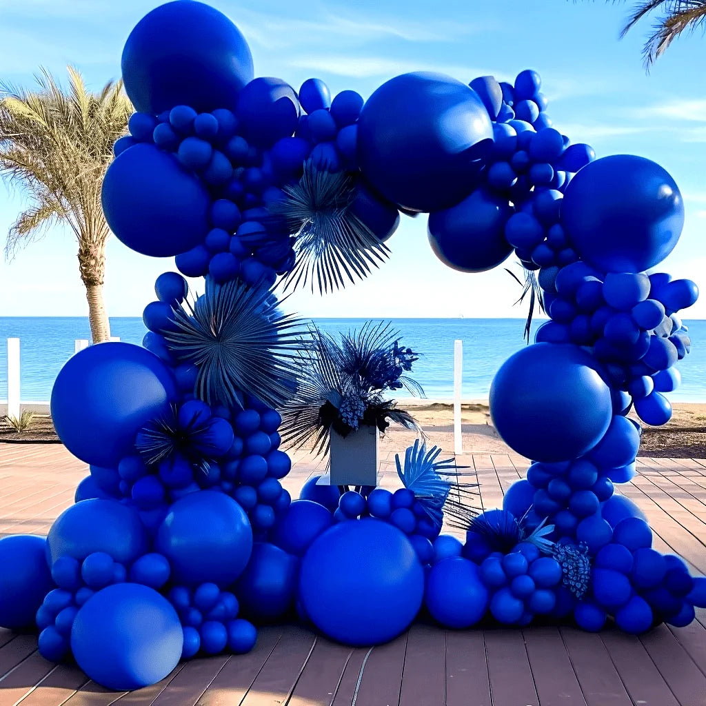 A large balloon arch garland made of royal blue balloons decorated with palm fronds on a wooden deck.