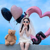 A young girl sits in front of a large pink heart balloon decoration with a black letter Q and a teddy bear.