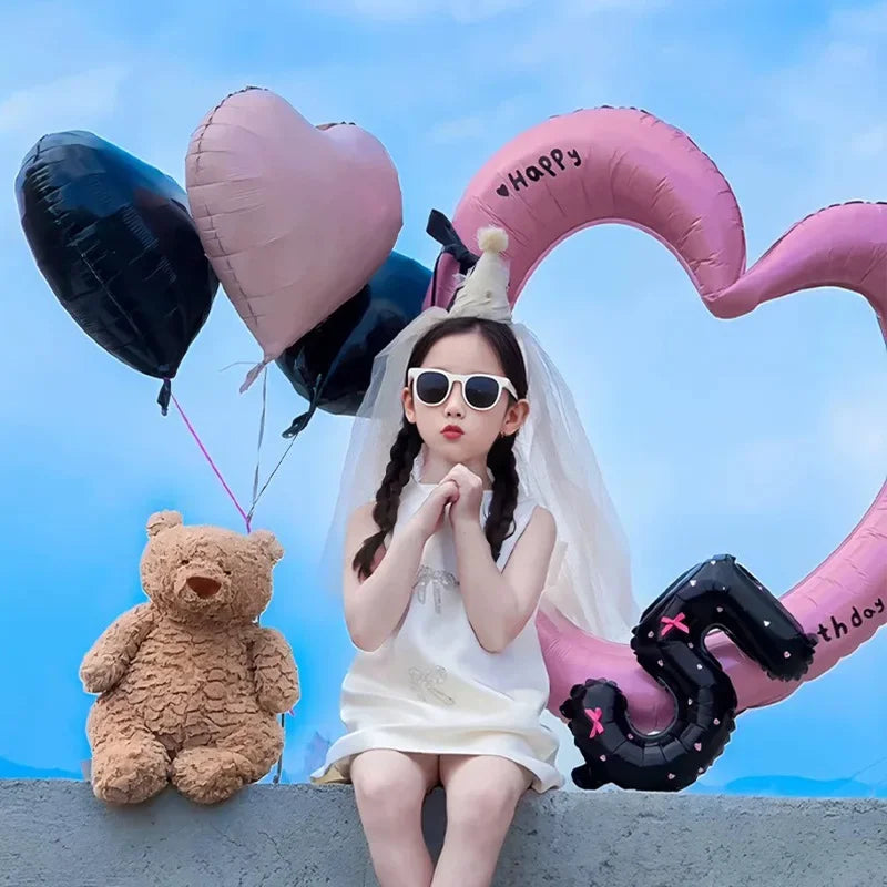 A young girl sits in front of a large pink heart balloon decoration with a black letter Q and a teddy bear.