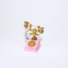 A vintage rotary desk phone miniature decor featuring brass-colored handset and base on a pink stand.