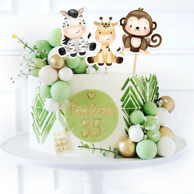 A white cake decorated with green and gold accents featuring jungle animal toppers like a zebra, giraffe, and monkey.