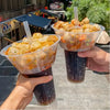 A hand holds a clear plastic cup filled with fried snacks and fruit in a dark colored drink cup.