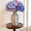 A hand-painted blue and white porcelain vase filled with purple hydrangeas, displayed on a wooden stand.