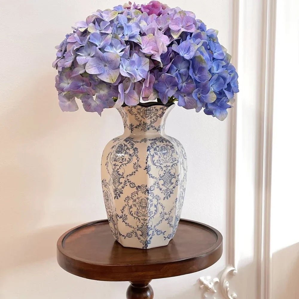 A hand-painted blue and white porcelain vase filled with purple hydrangeas, displayed on a wooden stand.