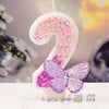 A white candle shaped like the number 2 decorated with pink and purple glitter, set against a blurred background.