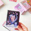 A hand holds a mini K-Pop photo card album binder featuring a heart-shaped photo and a small purple bear.