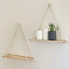 A wooden hanging shelf with hemp rope is mounted on a wall, displaying white candles and a potted green plant.