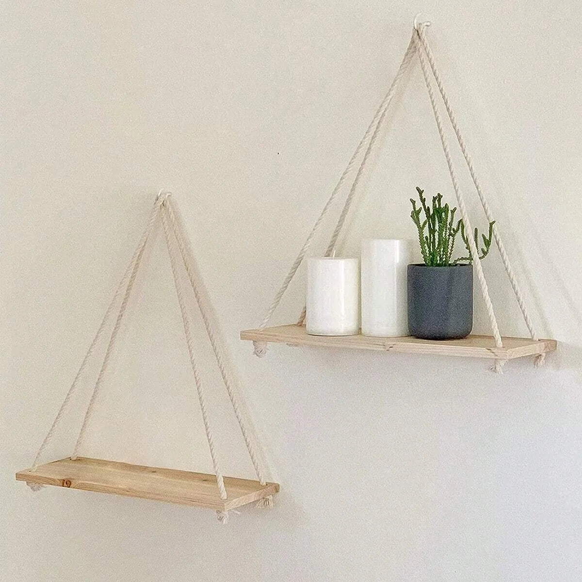 A wooden hanging shelf with hemp rope is mounted on a wall, displaying white candles and a potted green plant.