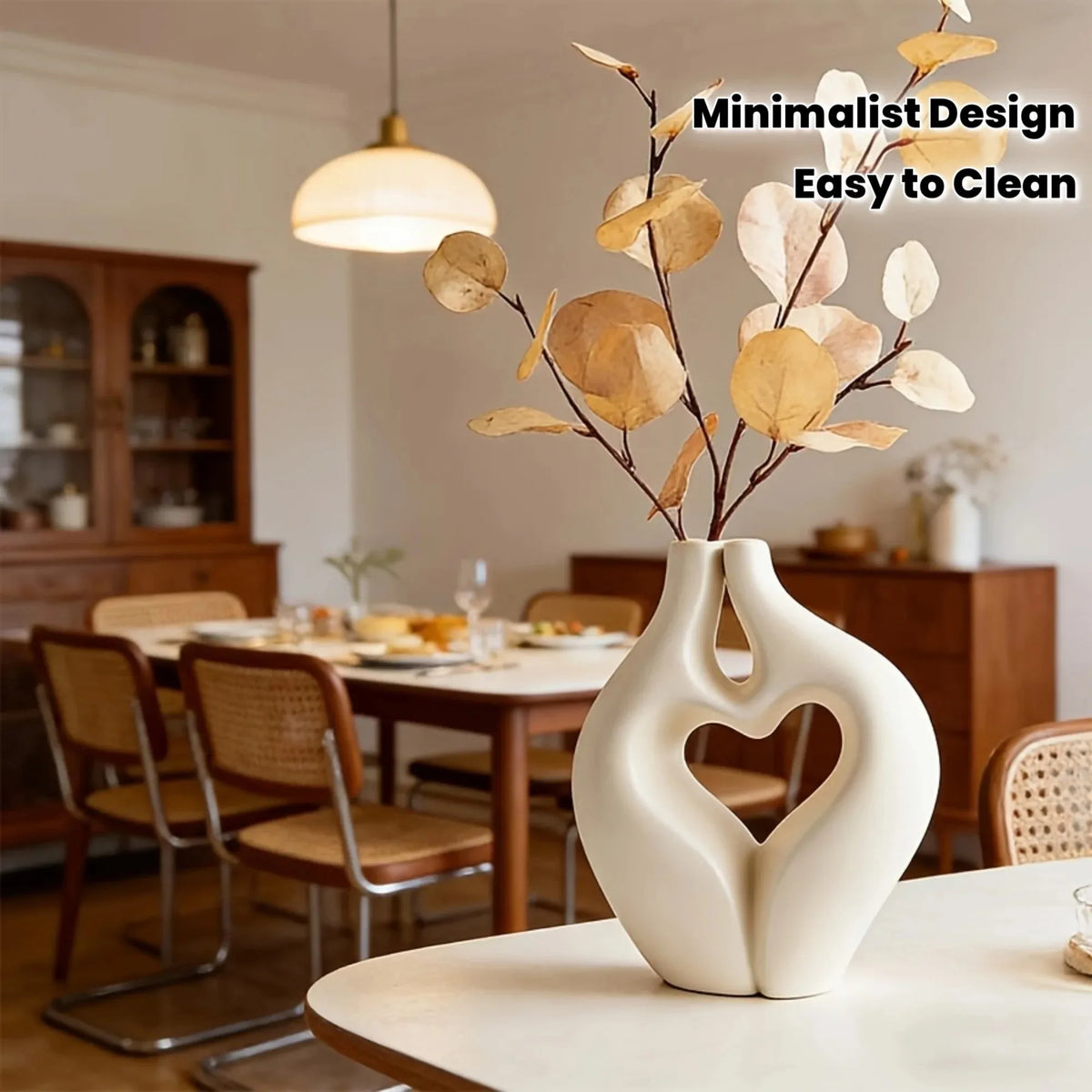 A white, modern heart-shaped vase holds dried botanical branches in a bright interior setting.
