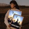 A person holds up a framed piece of Minecraft art featuring pixelated figures against a sunset background.