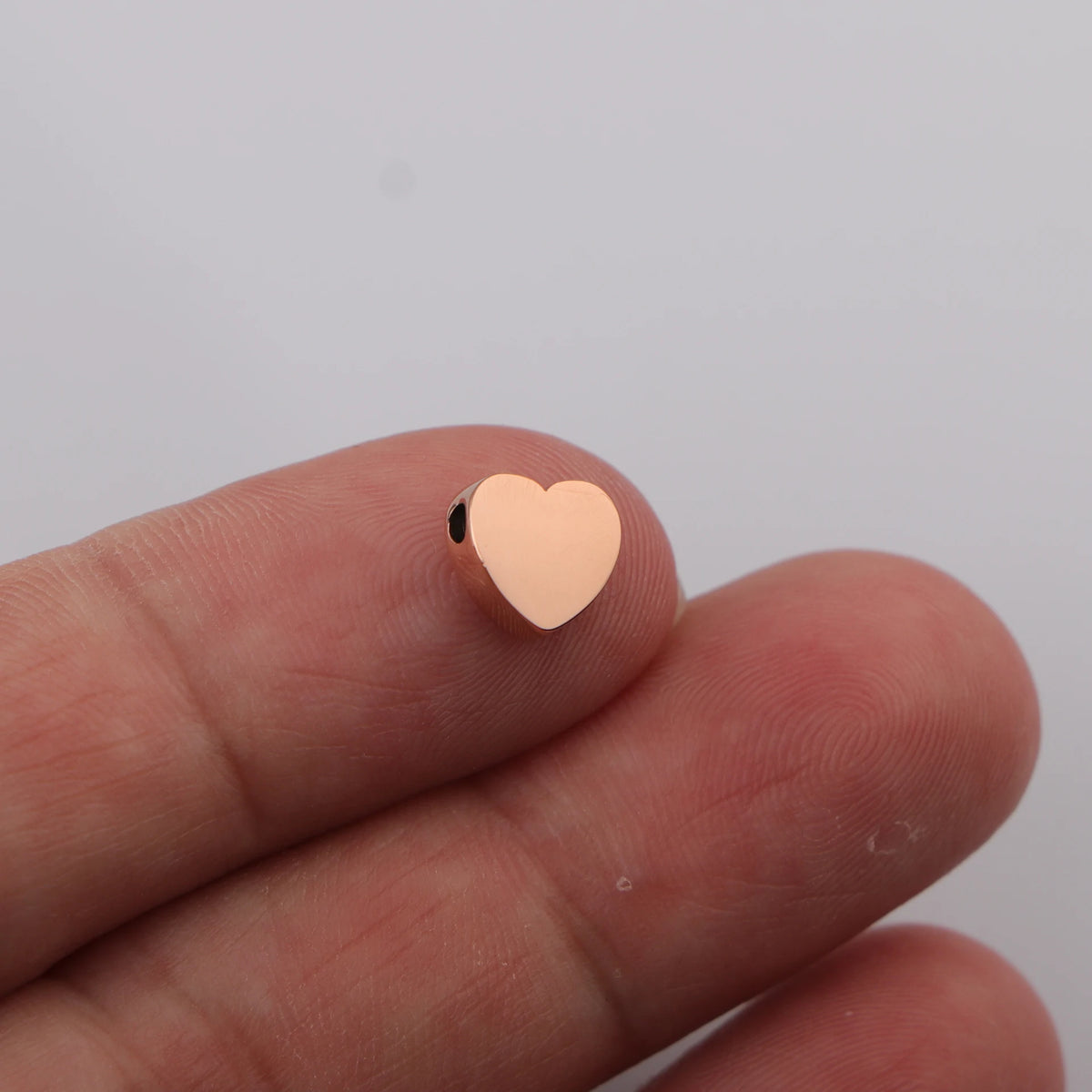 A hand holds a small, heart-shaped stainless steel bead charm against a white background.