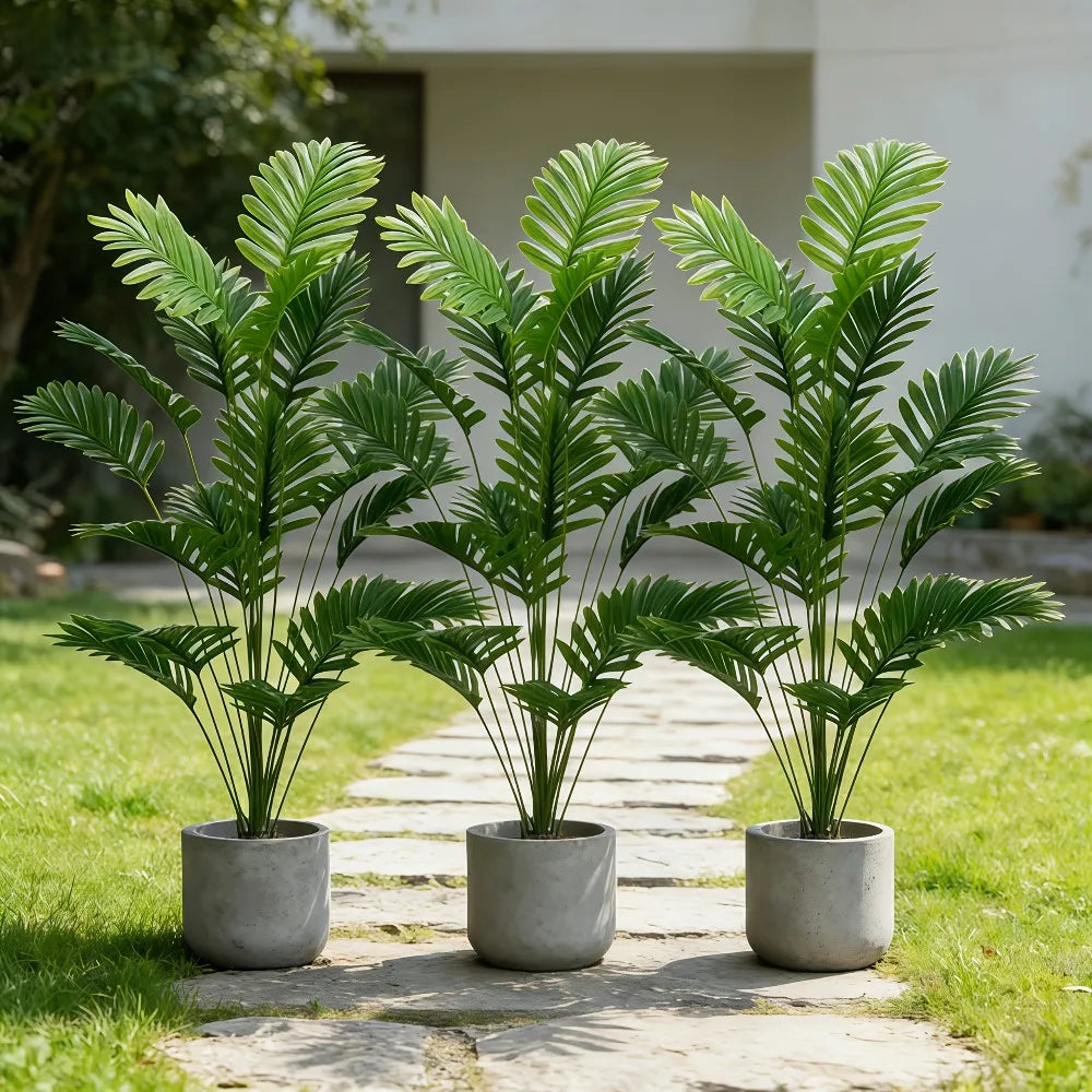 Three artificial palm trees with vibrant green monstera leaves planted in gray pots on a stone path.