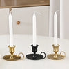 Three white taper candles in brass and black metal holders on a white table.