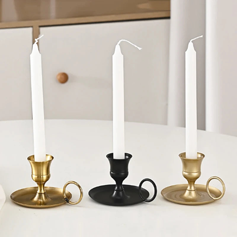 Three white taper candles in brass and black metal holders on a white table.