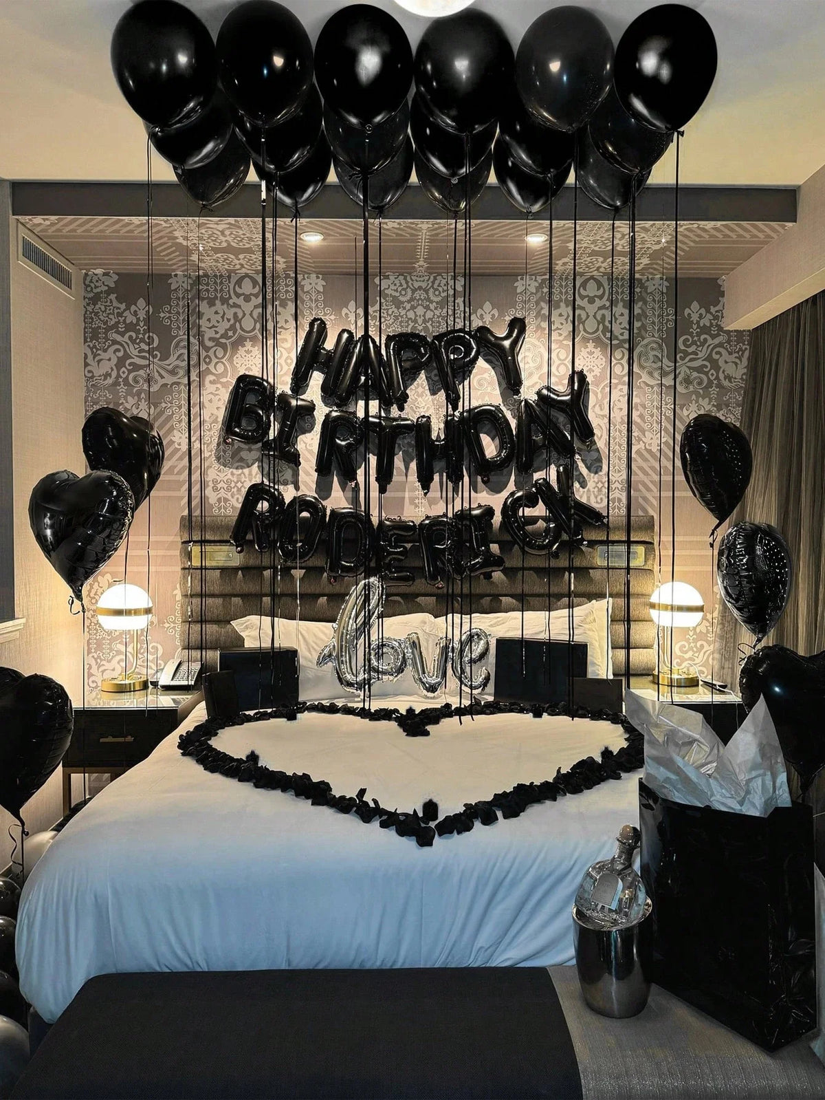 A bedroom decorated for a birthday party with black balloons and foil letters spelling 'HAPPY BIRTHDAY LOVE' above a bed.