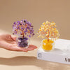 A hand holds a purple crystal money tree ornament and a yellow crystal money tree ornament on a white box.