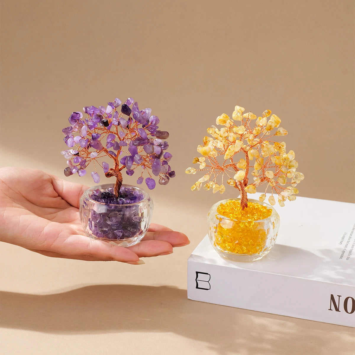 A hand holds a purple crystal money tree ornament and a yellow crystal money tree ornament on a white box.
