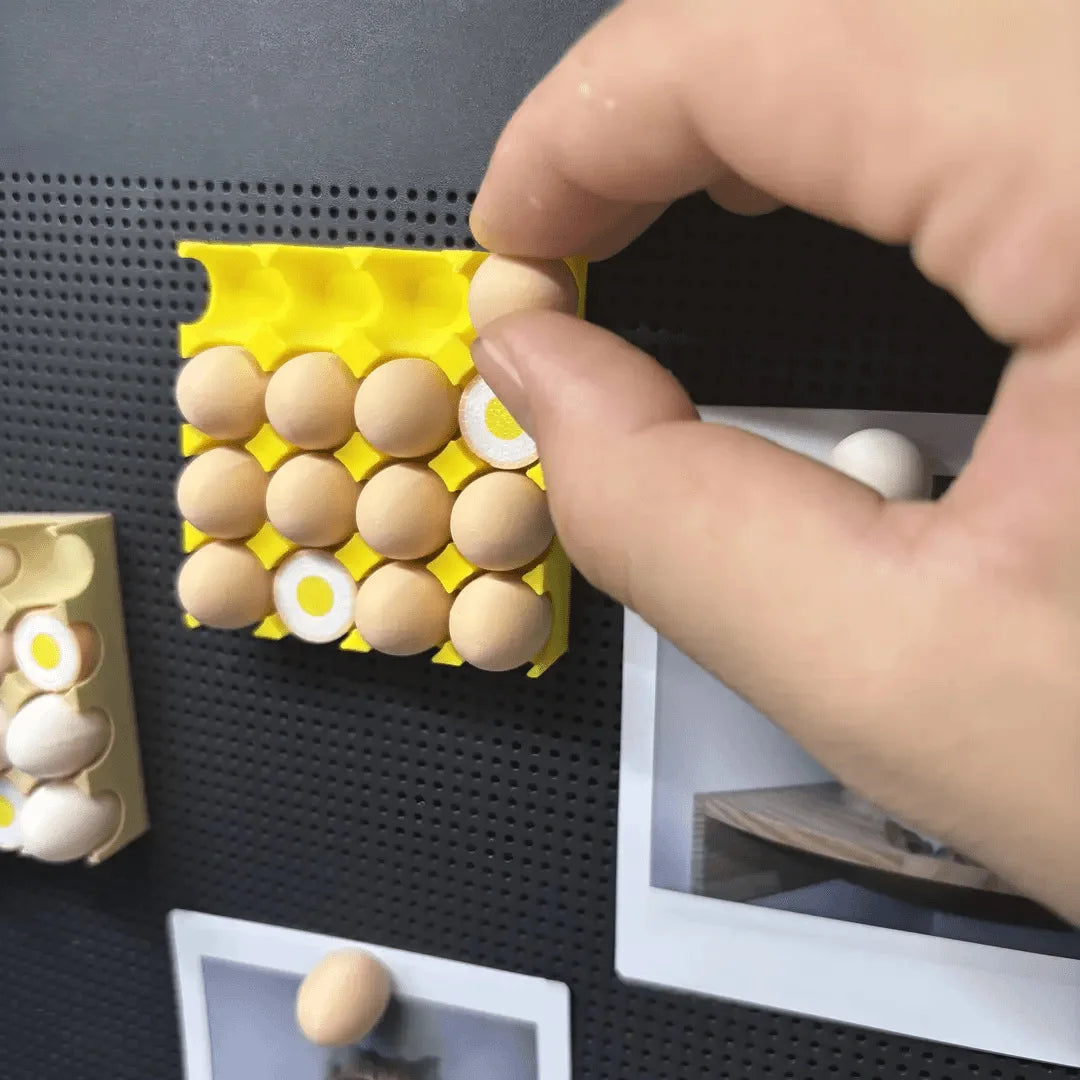 A hand is placing small, round, beige magnetic eggs into a yellow tray for fridge decor.