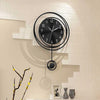 A modern black circular wall clock with Arabic numerals is mounted on a light-colored brick wall.