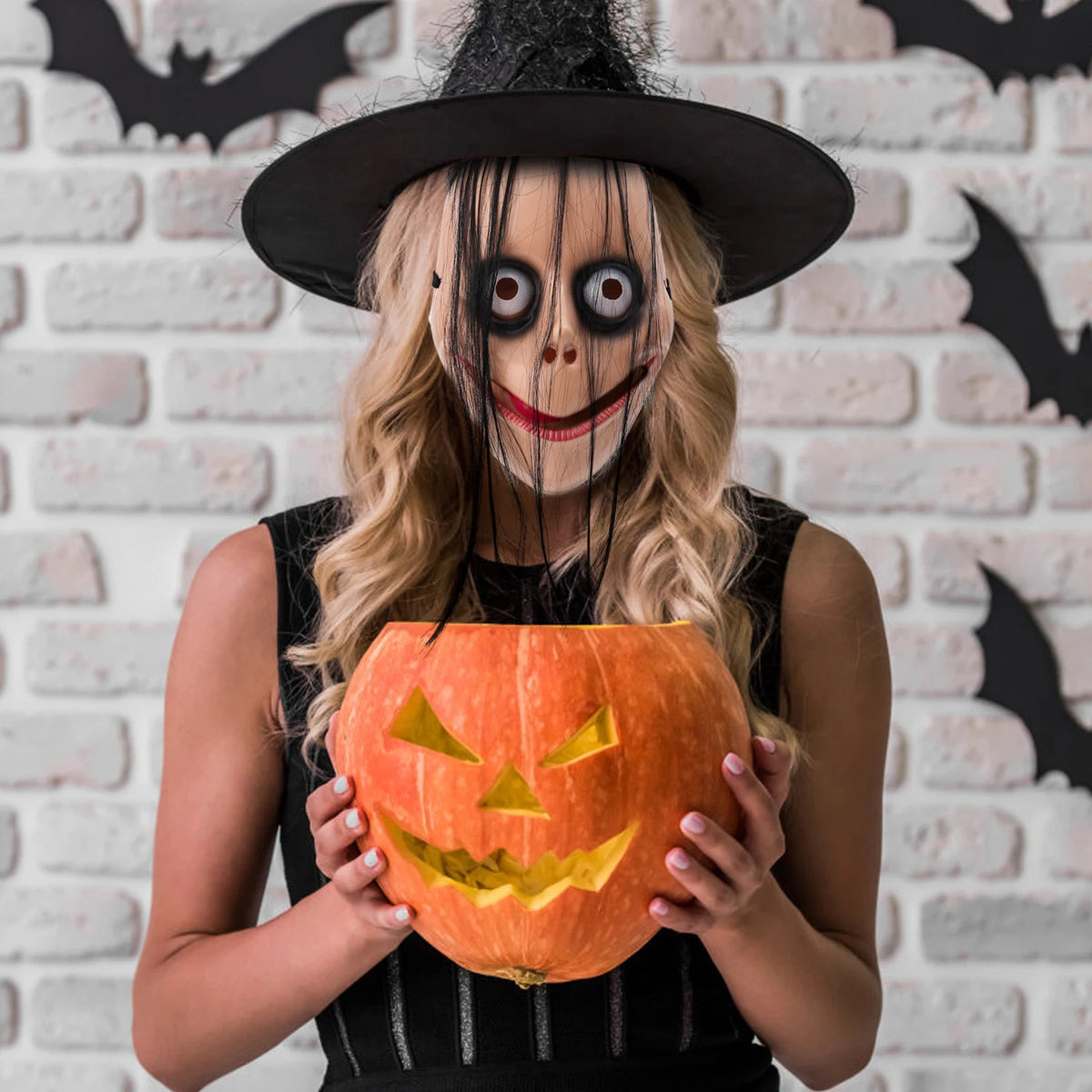 A woman wearing a creepy Halloween evil clown mask with exaggerated features holds an orange carved pumpkin.