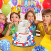 A birthday celebration featuring a multi-tiered white cake decorated with colorful foam balls and flowers, surrounded by happ