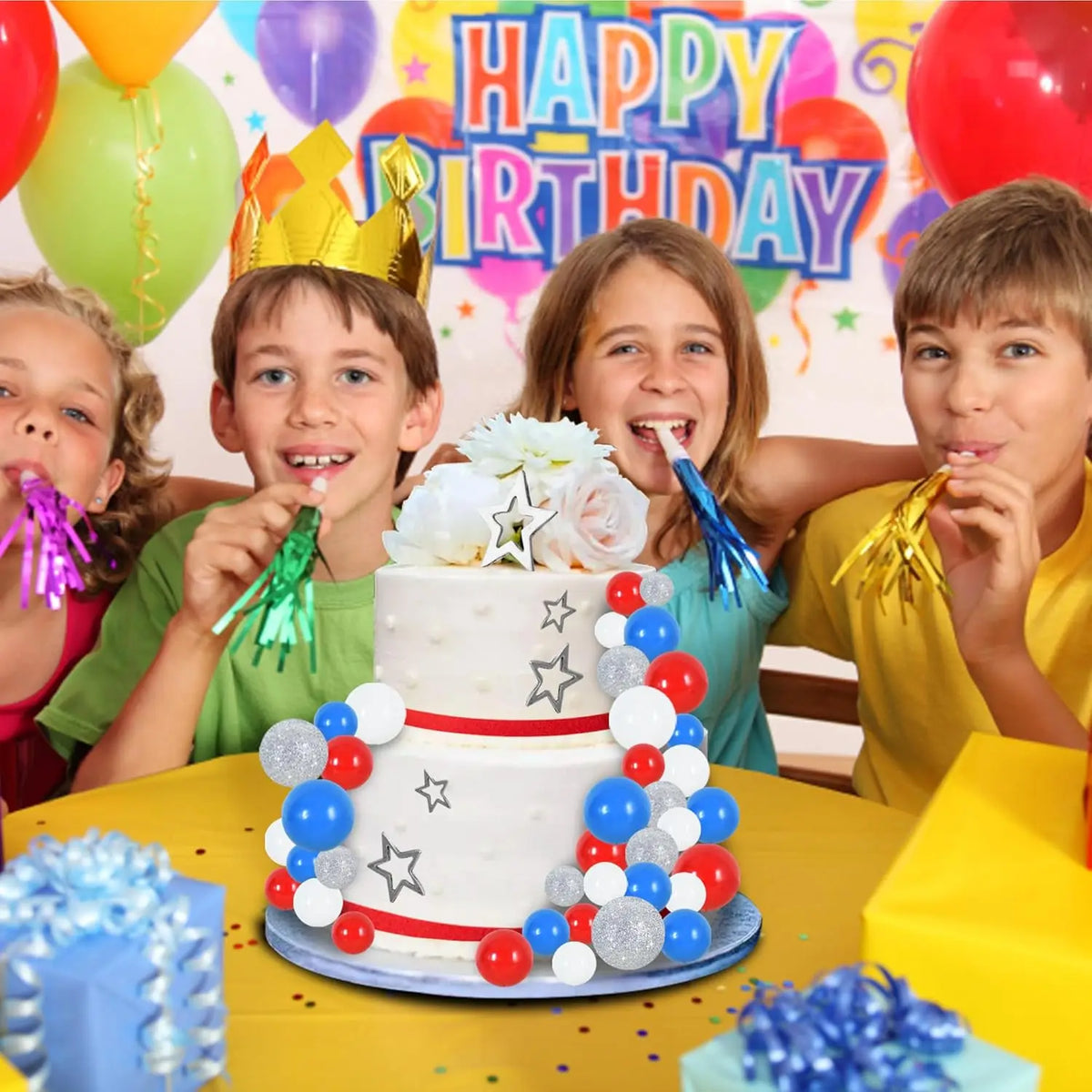 A birthday celebration featuring a multi-tiered white cake decorated with colorful foam balls and flowers, surrounded by happ