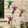 Four metal hummingbird wall decor figurines in vibrant colors mounted on a light-colored stone wall.