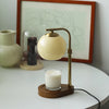 A brass aromatherapy wax melting lamp with a glass bulb sits on a wooden base next to a framed drawing.