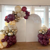 A balloon garland arch display featuring burgundy, gold, and white balloons against a white backdrop.