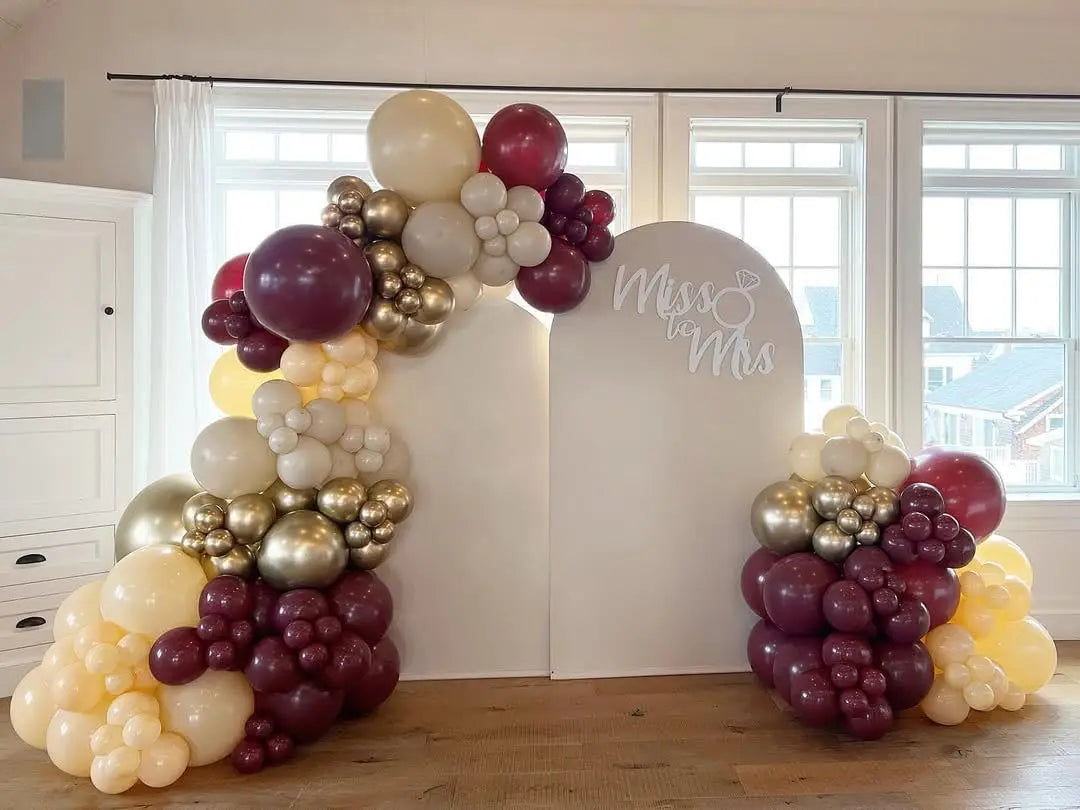 A balloon garland arch display featuring burgundy, gold, and white balloons against a white backdrop.
