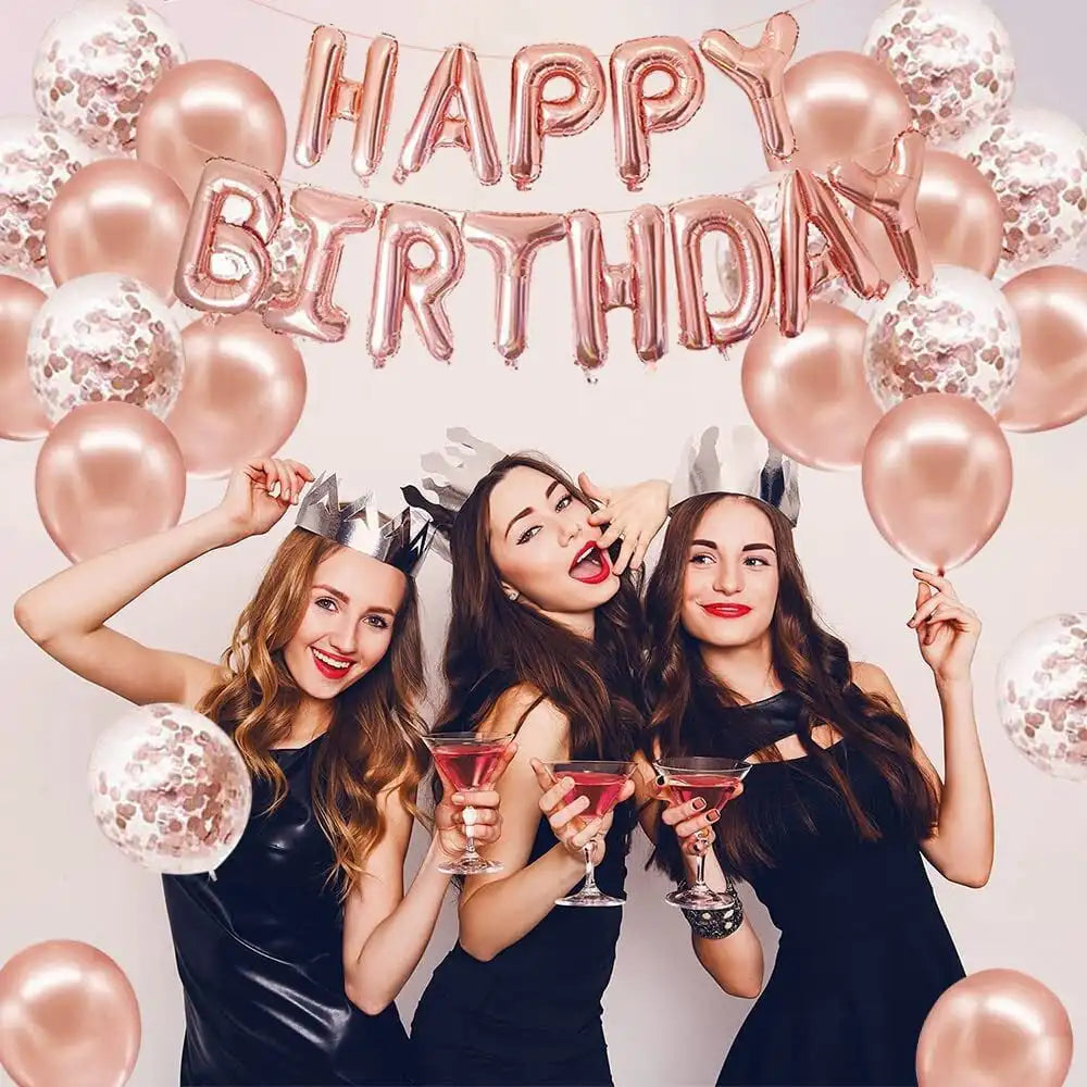 Three women in black dresses wear crowns and hold red drinks in front of a pink balloon garland.