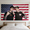 Two young men pose in front of an American flag tapestry hanging above a bed.