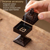 A hand holds a square incense accessory made of dark wood, designed to safely hold and isolate open incense flames.